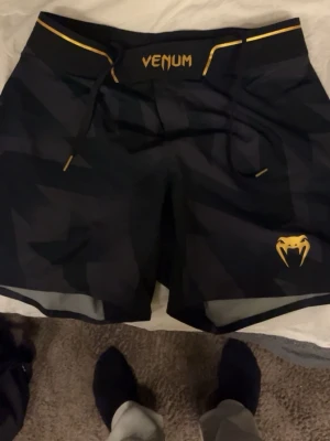 Svarta träningsshorts från Venum - Säljer ett par svarta träningsshorts från Venum med coolt geometriskt mönster och gul logotyp på benet. Shortsens midja har snörning och detaljer i gult. Perfekta för MMA, gym eller annan träning.