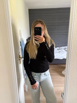 Svart ribbad långärmad tröja - Snygg svart ribbad långärmad tröja med smal passform och dekorativa silverknappar på ärmsluten. Perfekt att matcha med jeans eller kjol. Materialet ser ut att vara mjukt och stretchigt, vilket gör den bekväm att bära hela dagen.