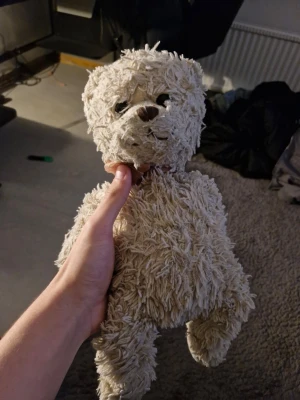 Beige mjukisdjur hund - Säljer ett gulligt beige mjukisdjur i form av en hund. Den har lurvig päls, svarta ögon och en brun nos. Perfekt som kramkompis eller dekoration i rummet.