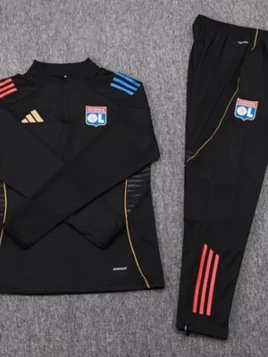 Svart träningsset Lyon Adidas - Snyggt svart träningsset från Adidas med Olympique Lyonnais-logga på både tröja och byxor. Tröjan har färgade ränder på axlarna, guldiga detaljer och halv dragkedja. Byxorna har röda ränder och smal passform. Perfekt för fotbollsträning eller gymmet.
