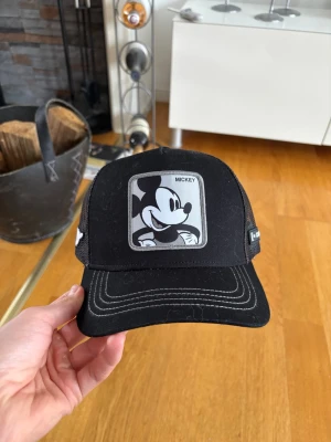 Svart truckerkeps med Mickey Mouse - Säljer en svart truckerkeps från Goorin Bros med ett stort Mickey Mouse-märke framtill och vit broderad Mickey-hand på sidan. Kepsen har mesh baktill för extra ventilation och justerbar snapback. Perfekt för dig som gillar Disney och streetwear. Aldrig andvänd!