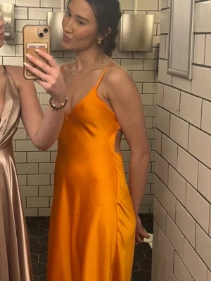 Orange långklänning i satin - Säljer en snygg orange långklänning med smala axelband och slätt, glansigt satinmaterial. Klänningen har en enkel och elegant design med figurnära passning och ärmlös modell. Perfekt för dig som vill lyfta upp en outfit 