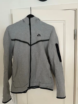 Grå Nike hoodie med dragkedja - Snygg grå hoodie från Nike med hel dragkedja framtill och svarta detaljer. Har huva, två fickor fram och en extra ficka med dragkedja på ärmen. Tillverkad i mjukt material, perfekt för chill eller träning. Nike-logga på bröstet och cool design med svarta linjer.