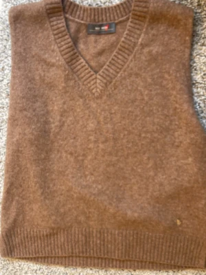 Brun ullväst från Mos Mosh - Snygg brun väst från Mos Mosh i mjuk ull med v-ringad hals och ribbade kanter. Perfekt att slänga över en skjorta för en chill och stilren look. Klassisk design som funkar till mycket.