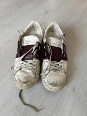 Valentino vita och vinröda sneakers - Snygga sneakers från Valentino i vitt läder med vinröda detaljer över mitten. Klassisk låg modell med vita snören och gummisula. Märkeslogga på plösen och coola nitar baktill på sulan. Perfekta för dig som gillar streetstyle och designer. Storlek 36-37 skulle jag säga. OBS!!! Priset är rörligt