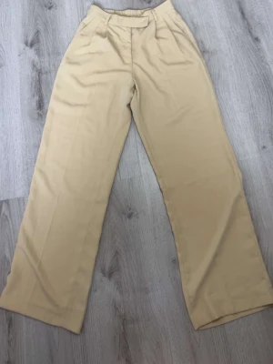 Beige vida chinos med hög midja - Snygga beige chinos med hög midja och vida ben. Byxorna har klassisk look med pressveck framtill och elastisk resår bak i midjan för extra komfort. Perfekta för en clean och avslappnad stil. Materialet känns mjukt och följsamt. Skriv om en bild hur den är på