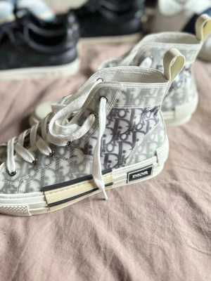 Dior B23 High Top sneakers - Snygga Dior B23 High Top sneakers med genomskinlig mesh och det ikoniska Dior Oblique-mönstret i grått och vitt. Skorna har vita snören, gummisula och detaljer i vitt och beige. Perfekt för dig som vill sticka ut med exklusiva och trendiga sneakers. (Finns ingen låda till) 