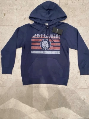 Mörkblå Saint Laurent hoodie - Mörkblå hoodie från Saint Laurent med tryck i vitt och rött framtill. Klassisk känguruficka och justerbar huva med snören. Tillverkad i mjukt bomullsmaterial för en skön och avslappnad look. Perfekt för dig som vill ha en stilren och trendig hoodie.