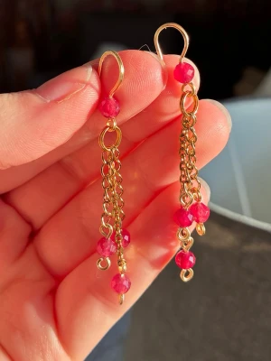 Guldörhängen med röda pärlor - Säljer ett par snygga örhängen i guldfärgad metall med flera kedjor och röda pärlor. Örhängena har en hängande design med små, facetterade pärlor som ger ett coolt och färgstarkt intryck. Perfekt för dig som gillar unika accessoarer.