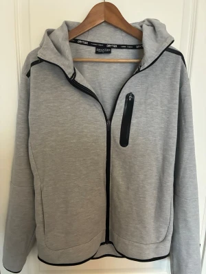 Grå hoodie från Drytex med dragkedja - Snygg grå hoodie från Drytex med hel dragkedja framtill och svart dragkedjeficka på bröstet. Hoodien har svarta detaljer längs dragkedjor och axlar samt en skön huva. Perfekt för en avslappnad och sportig stil.
