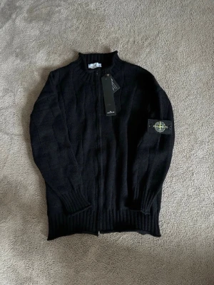 Svart stickad ziptröja Stone Island - Svart stickad ziptröja från Stone Island med ribbad ståkrage och ärmslut. Tröjan har dragkedja framtill och den klassiska Stone Island-loggan på vänster ärm. Perfekt för lager-på-lager och snygg till jeans.
