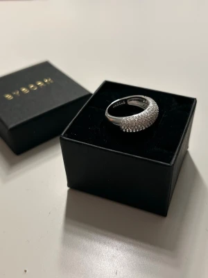 Silverring med stenar från Byborn - Stilren silverfärgad ring från Byborn med glittrande små stenar tätt placerade på ovansidan. Ringen har en rundad form och levereras i original ask. Perfekt för dig som gillar eleganta accessoarer med extra bling. Aldrig använd 