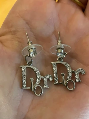 Silverfärgade Dior örhängen (ej äkta) - Säljer ett par coola silverfärgade örhängen från Dior med gnistrande stenar på bokstäverna. (ej äkta) Örhängena har en unik design där Dior-loggan är i fokus och är tillverkade i metall med stenar som ger extra bling.
