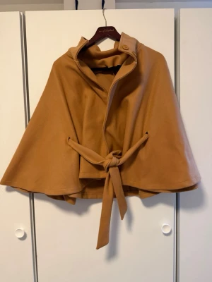 Camelbrun cape med knytband - Stilren camelbrun cape med knytband i midjan och hög krage med knappdetalj. Capen har en vid, svepande form och är tillverkad i ett mjukt tyg som ger en elegant look. Perfekt för lager på lager och trendiga outfits.