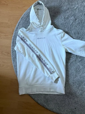 Gant hoodie - Snygg vit hoodie från GANT med broderad logga på bröstet och  band med GANT-tryck längs ärmarna. Perfekt för en clean och stilren look. 