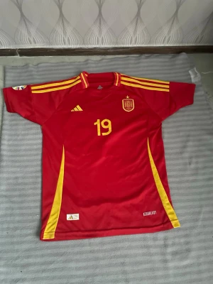 Spansk landslagströja Adidas nr 19 - Röd fotbollströja från Adidas med gula detaljer, nummer 19 och namn Lamine Yamal på ryggen. Officiell spansk landslagströja med klubbmärke på bröstet och Adidas-logga. Korta ärmar och ventilerande material, perfekt för match eller träning.