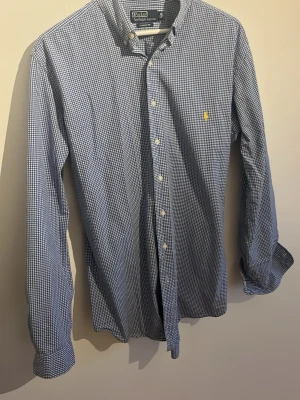 Blårutig skjorta från Polo Ralph Lauren - Säljer en klassisk blårutig skjorta från Polo Ralph Lauren med broderad gul logga på bröstet. Skjortan har lång ärm, knappar framtill och är gjord i mjuk bomull. Perfekt för dig som gillar tidlös stil och snygga detaljer. OBS stor i storlek!