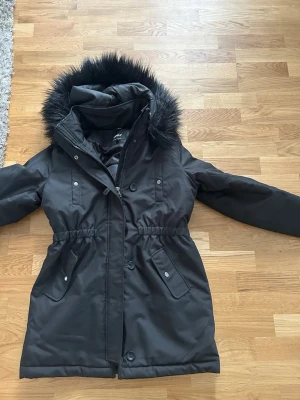 Svart parkasjacka med fuskpäls - Snygg svart parkasjacka från ONLY med avtagbar huva och fuskpäls. Jackan har dragkedja och knappar framtill, två stora fickor med lock och markerad midja för en schysst siluett. Perfekt för kalla dagar och riktigt varm.