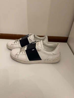 Vita Valentino sneakers med nitar - Säljer ett par vita sneakers från Valentino med svart band över vristen och coola nitar runt hälen. Skorna har klassisk låg profil, vita snören och är tillverkade i läder. Perfekta för dig som gillar en stilren men edgy look.