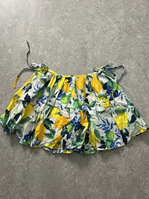 Kort kjol med citronmönster och knyt - Säljer en färgglad kort kjol med citron- och bladmönster i gult, grönt och blått. Kjolen har volang och knytband i midjan för en söt och somrig look. Perfekt för dig som vill sticka ut med en unik stil!
