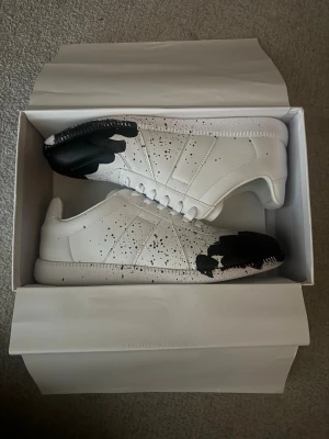 Maison Margiela Replica sneakers svart/vit - Maison Margiela Replica sneakers i vitt läder med svart färgstänk och svart tå. Klassisk låg modell med vita skosnören och unika detaljer. Snygg och modern design som sticker ut med sin konstnärliga touch.