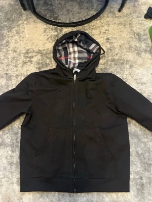 Svart Burberry hoodie med dragkedja - Säljer en svart hoodie från Burberry med dragkedja framtill och klassiskt rutigt foder i huvan. Hoodien har två fickor och en diskret broderad Burberry-logga på bröstet. Perfekt för dig som gillar stilrena plagg med exklusiva detaljer.