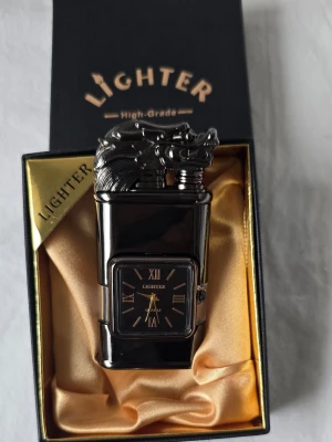 Svart tändare med drakmotiv och klocka,fler modellen  - Unik svart tändare med drakmotiv på toppen och inbyggd analog klocka med romerska siffror. Tändaren har en blank metallfinish och ett fönster för gasnivå. Levereras i elegant presentask med guldfärgad insida.