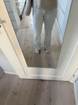 Grå mjukisbyxor med dragsko - Snygga ljusgrå mjukisbyxor med resår och dragsko i midjan. Byxorna har raka ben och en broderad detalj med bokstaven 'M' vid höften. Perfekta för en chill och avslappnad stil. Tillverkade i mjukt och bekvämt material.