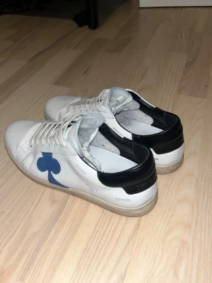 Vita sneakers från The Gallerist - Stilrena vita sneakers från The Gallerist med svart häl och blå klubb-symbol på sidan. Skorna har rund tå, snörning och platt sula. Tillverkade i skinn och levereras med originalkartong och dustbag.