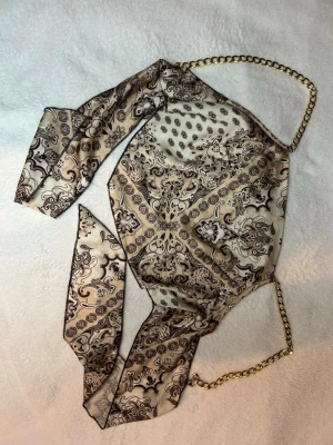 Beige mönstrad scarf-topp med kedja - OANVÄND Scarf-topp i beige och svart med paisleymönster och guldfärgade kedjor som axelband. Tunn och glansig satinliknande känsla, perfekt för att sticka ut. Knytning i ryggen för justerbar passform.