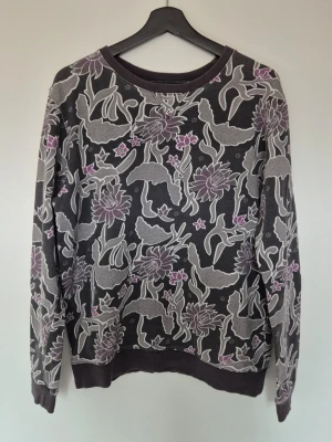 Contemporary Reflections Svart mönstrad långärmad topp - Cool svart långärmad topp med unikt mönster i grått och lila, med blommor och abstrakta former. Rund halsringning och ribbade muddar vid ärmslut och nederkant. Perfekt för dig som vill sticka ut med en kreativ och konstnärlig stil.