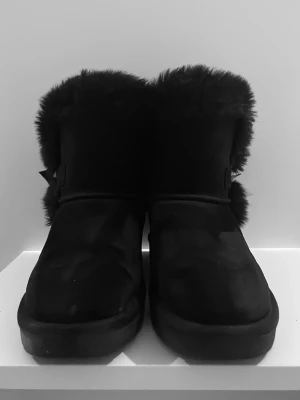 Svarta och varma boots🖤 - Mysiga svarta uggs med fluffig fuskpäls runt skaftet och en söt rosett på sidan. Uggsen har en mjuk ovandel och tjock sula, perfekta för kalla dagar hemma. Klassisk slip-in modell som ger extra värme och stil.