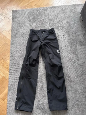 Svarta täckbyxor från Haglöfs - Säljer ett par svarta täckbyxor från Haglöfs med raka ben och elastisk midja. Byxorna har dragkedjor längs benen och är tillverkade i ett slitstarkt, vattenavvisande material. Perfekta för kalla dagar utomhus eller till vintersport.