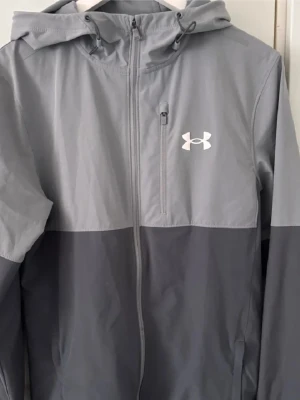Grå vindjacka från Under Armour - Snygg tvåtonad grå vindjacka från Under Armour med huva och hel dragkedja. Jackan har en bröstficka med dragkedja och vitt logotryck på bröstet. Tillverkad i lätt och vindtätt material, perfekt för blåsiga dagar.