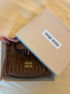Handväska Miu Miu - Säljer en brun quiltad handväska från Miu Miu i mjukt läder med guldfärgade detaljer och logotyp framtill. Väskan har en rundad form och kort handtag, perfekt för att bära i handen. Kommer med original dustbag och box.