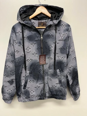 Grå Louis Vuitton vindjacka med monogram - Snygg grå vindjacka från Louis Vuitton med ikoniskt monogrammönster över hela jackan. Jackan har huva med svarta dragsnören, hel dragkedja framtill och två fickor. Materialet är lätt och passar perfekt för blåsiga dagar.