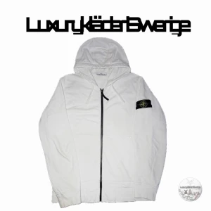  Stone Island Vit hoodie  - ÄKTAs Snygg vit hoodie från Stone Island med dragkedja framtill och klassisk logotyp-patch på vänster ärm. Hoodien har huva med dragsko och är tillverkad i mjukt bomullsmaterial. Perfekt för en clean och stilren look.