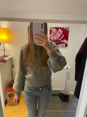 Grå stickad tröja med ballongärm - Mysig grå stickad tröja med ballongärmar och ribbade muddar vid ärmslut och midja. Tröjan har en klassisk rund halsringning och är tillverkad i ett mjukt garn som ger en avslappnad look. 