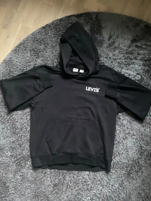 Svart hoodie från Levi's med huva - Säljer en svart hoodie från Levi's i relaxed fit med korta ärmar och stor huva. Klassisk känguruficka framtill och Levi's-logga tryckt på bröstet. Tillverkad i mjukt material, perfekt för en chill och avslappnad stil. Helt ny!!!