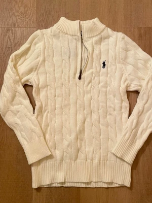 Vit kabelstickad tröja Polo Ralph Lauren - Kabelstickad vit tröja från Polo Ralph Lauren med halv dragkedja och hög krage. Tröjan har lång ärm, ribbade muddar och den klassiska mörkblå logotypen broderad på bröstet. Perfekt för dig som gillar stilren och tidlös design.