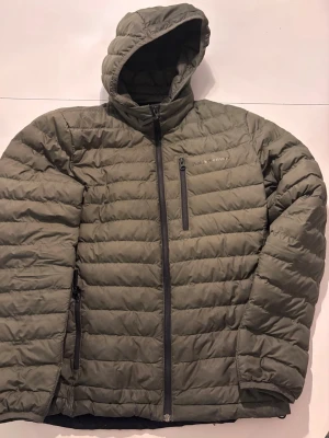 Peak Performance grön jacka  - Grön Peak Performance jacka med huva och dragkedja framtill, herrmodell strl S. Modell M Rivel Liner Jacket.  Vattenavstötande återvunnen polyester. Dragkedja, två framfickor med dragkedjor. Diskret logga på bröstet, dragsko vid plaggslut och små, elastiska muddar i ärmar och luva. Luvan är fäst med dragkedja och kan tas av. Bara höra av sig, pris ej hugget i sten!!