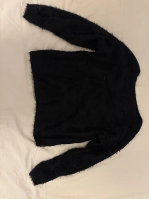 Svart fluffig stickad tröja - Mysig svart stickad tröja med fluffig, lurvig textur. Tröjan har rund halsringning och långa ärmar. Perfekt för dig som gillar en enkel men ändå trendig look. Materialet känns mjukt och ger en cool, avslappnad vibe.