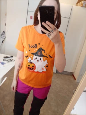 Orange Halloween t-shirt med spöke - Säljer en orange t-shirt med ett Halloweenmotiv på framsidan. Trycket visar ett gulligt spöke med häxhatt, fladdermöss, stjärnor och en pumpa. T-shirten har rund hals och korta ärmar, perfekt för dig som gillar roliga och färgglada plagg.