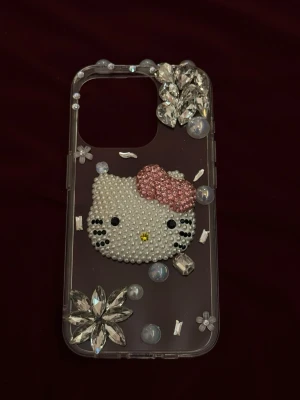 Hello Kitty mobilskal med pärlor & strass iPhone 15 PRO  - Unikt genomskinligt mobilskal dekorerat med en stor Hello Kitty i pärlor, rosa rosett och massor av glittrande strasstenar och blommor. Skalets material är plast och dekorationerna ger en riktigt söt och glamorös känsla. Perfekt för dig som vill sticka ut och älskar kawaii-stil. iPhone 15 PRO. 