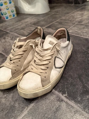 Philippe model skor  - Snygga vita sneakers med beige mockadetaljer och svarta detaljer vid hälen. Klassisk låg modell med vita snören och diskret logga på sidan. Perfekta för en avslappnad streetstil och passar till det mesta i garderoben.