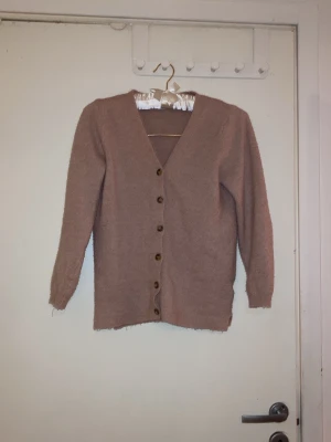 Beige kofta med knappar - Mysig beige kofta med v-ringning och bruna knappar framtill. Långärmad och rak modell, perfekt att slänga över en t-shirt eller topp. Koftan är stickad i ett mjukt material och har ribbade muddar vid ärmslut och nederkant.