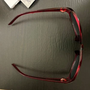 Röda ovala solglasögon med stenar - Snygga solglasögon med oval form och transparent röd båge. Bågarna har små glittriga stenar längs ovansidan för extra detalj. Glasen är mörka och ger en cool kontrast till den röda färgen. Perfekta för dig som vill sticka ut i solen.