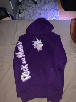 Lila Rick and Morty hoodie - Säljer en lila hoodie med Rick and Morty-tryck på ärmen och en tecknad Rick på bröstet. Hoodien har huva med dragsko och en stor magficka. Perfekt för dig som gillar serien och vill sticka ut med en färgglad och bekväm stil.