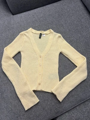 Gul ribbstickad kofta från H&M - Snygg ljusgul ribbstickad kofta från H&M med v-ringning och knappar framtill. Modellen är croppad med långa ärmar och mjukt, stretchigt material. Perfekt att slänga över en topp för en chill look.