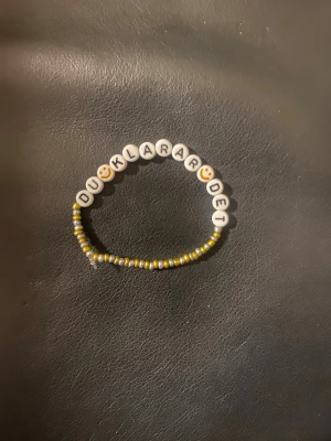 Pärlarmband med text och smileys - Fint armband med vita bokstavspärlor som bildar texten 'DU KLARAR DET' och två smiley-pärlor. Resten av armbandet består av små pärlor i gult och silver. Perfekt accessoar för att peppa dig själv eller någon annan.